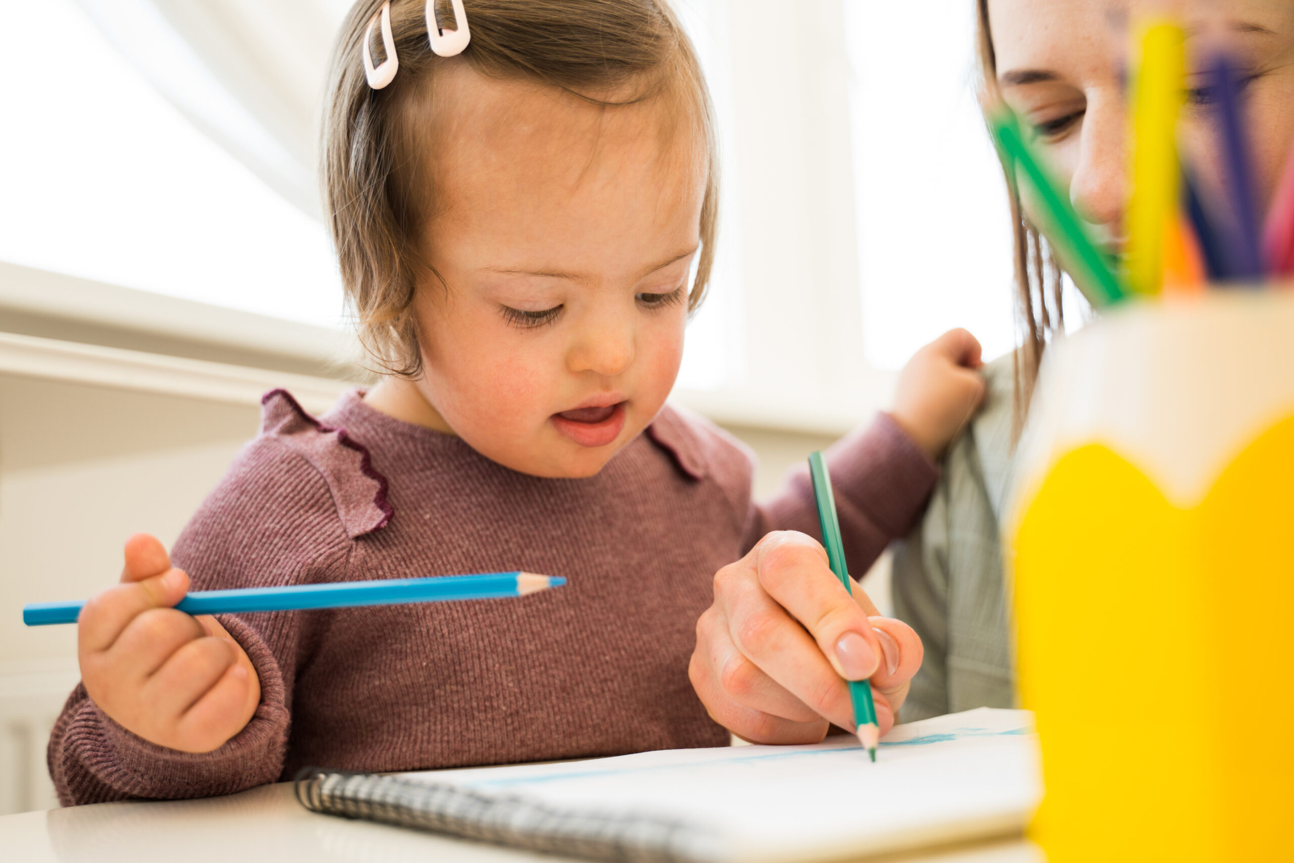Cute girl with down syndrome drawing and sitting over a blank sheet of paper at the table. Girl spending time with her mother and sharing positive emotions with her