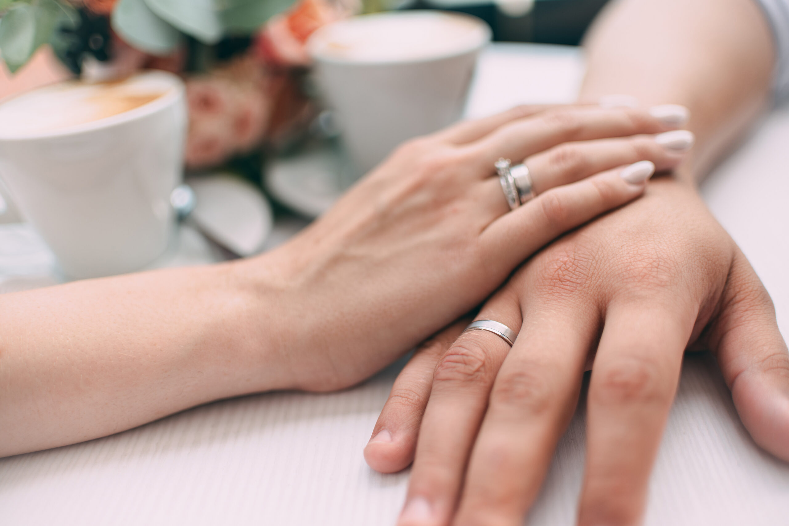 The bride and groom hold hands, holding expensive wedding rings with white gold, on the background of a wedding bouquet and two cups of coffee on a white table
